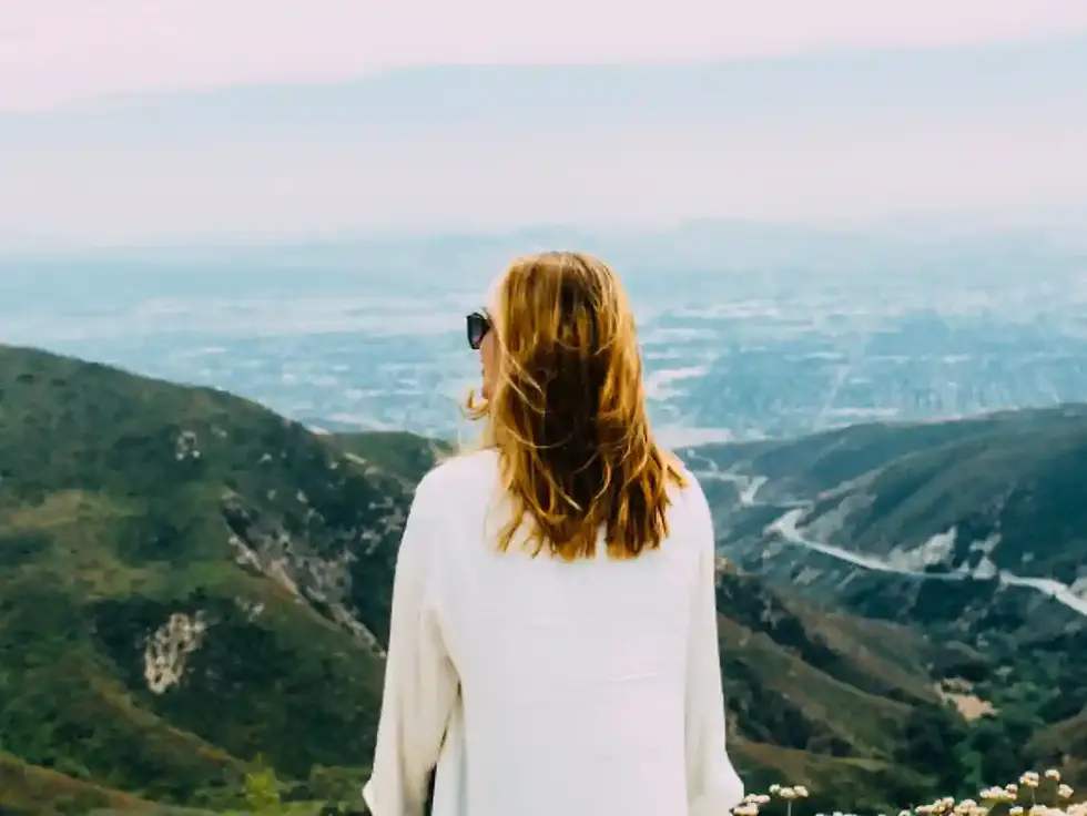 Woman in sunglasses gazes over scenic green mountains and cityscape. She's wearing a white shirt. Sky is light pink, creating a serene mood.