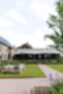 Llantilio House features a beautiful garden with manicured lawns and rustic seating, leading up to a charming brick building under a wide awning, surrounded by lush greenery and vibrant flowers under a clear sky.
