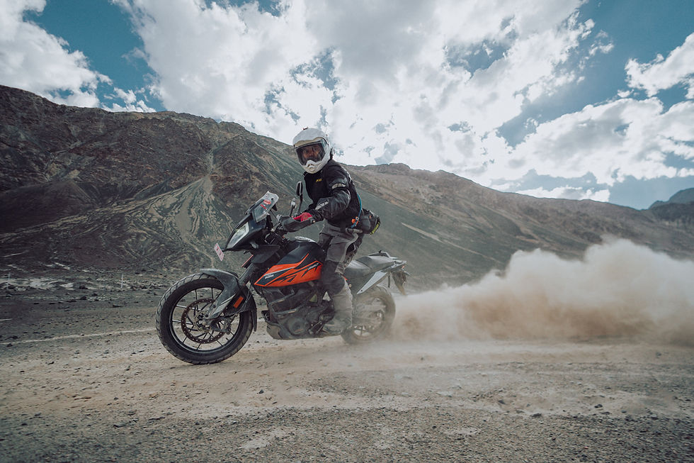 Motorcyclist riding on dirt road, kicking up dust, mountains in the background.
