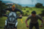 A person on motorcycle with another person standing beside in field