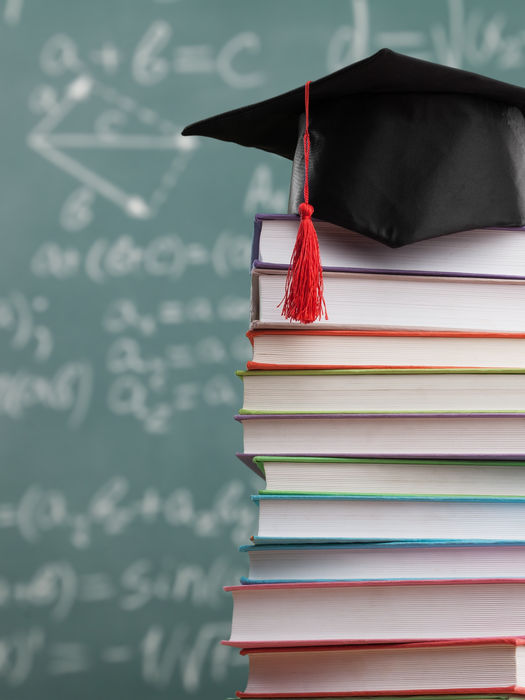 Stacks of college books in front of a caulk board with a graduation cap on top