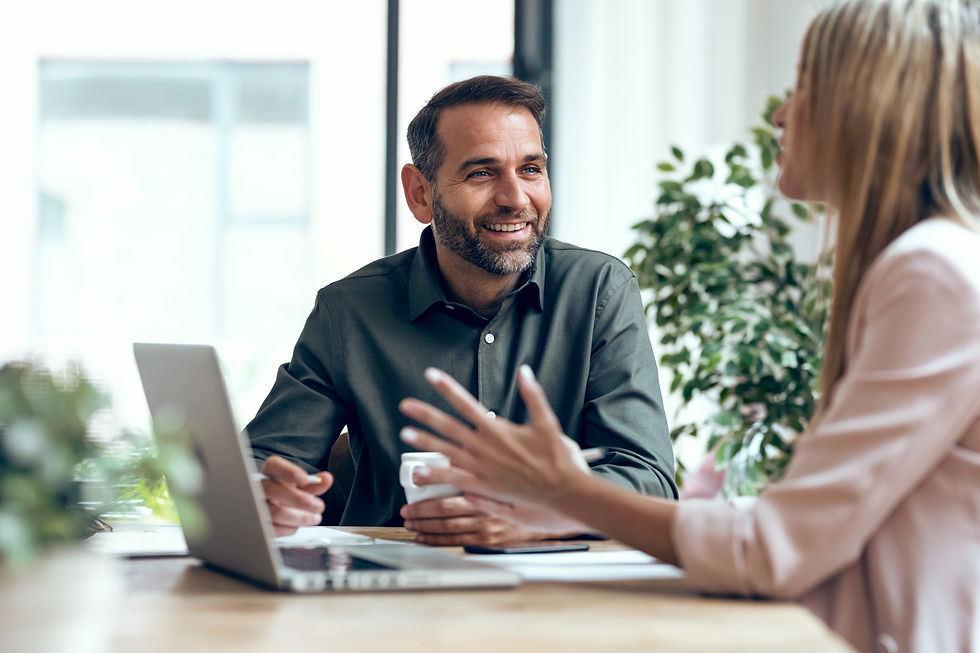 Smiling man and woman talking in an office with a laptop and papers on the table, greenery in the background, conveying a cheerful mood.