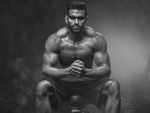 Muscular man sitting, looking forward with clasped hands in a dramatic pose.