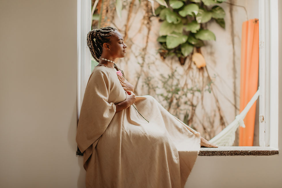A woman in a beige dress is sitting on a windowsill. Her eyes are closed and she is smiling. She has one hand on her heart. Both her hands are holding a single rose.