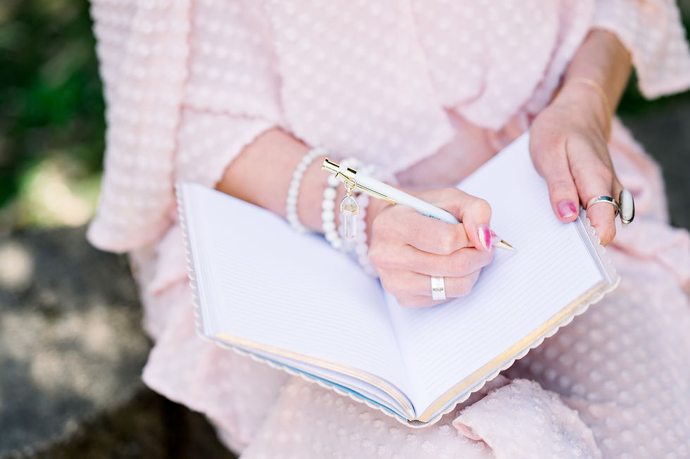 A woman is wearing a pink dress and crystal bracelets. Her hands are visible and she's writing in her journal.