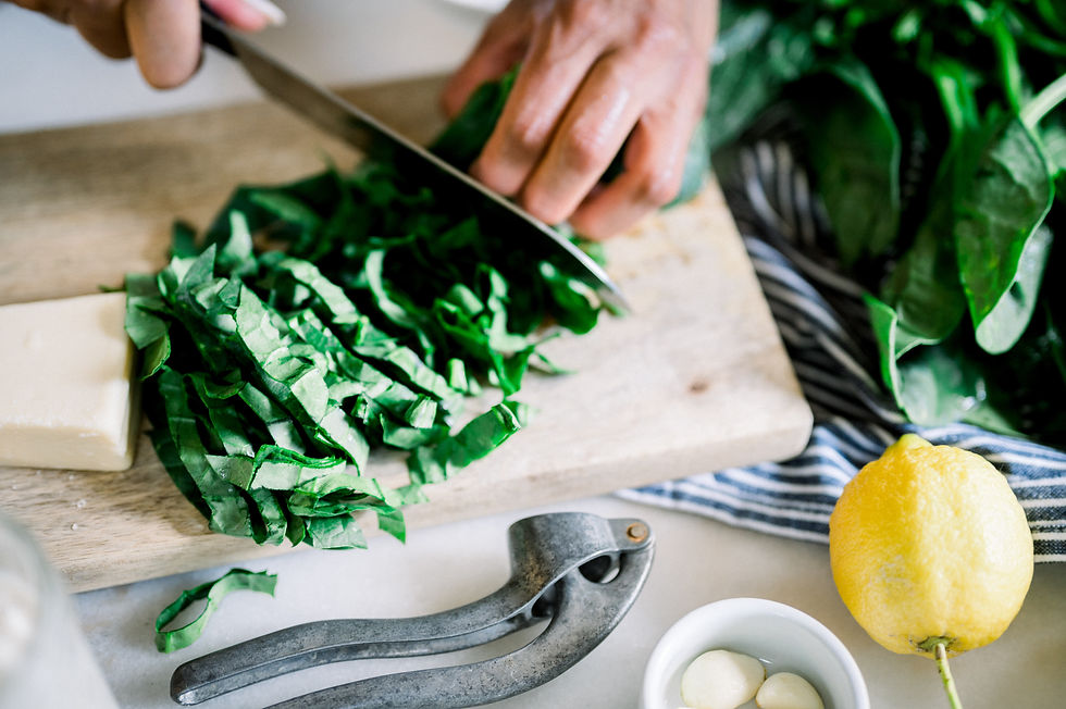 Hands slicing green leaves on a wooden board, next to a cheese block, lemon, garlic, and metal press. Striped cloth in the background.