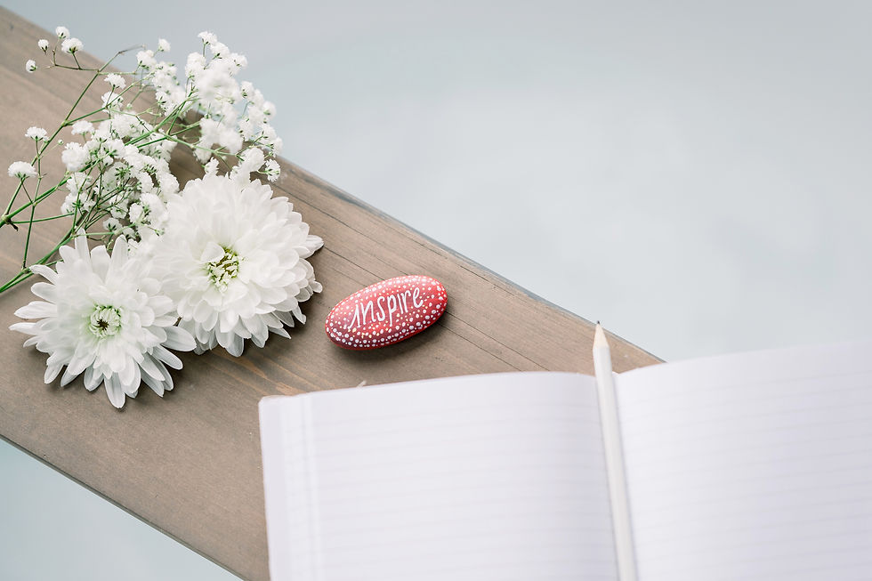 White flowers, a red stone with the word inspire, and an open journal sit on top of a table.