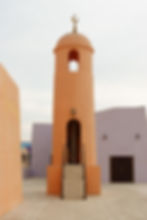 Tall orange tower with crescent on top, flanked by staircase. Background: pale purple walls, mural with eyes. Calm, overcast sky.