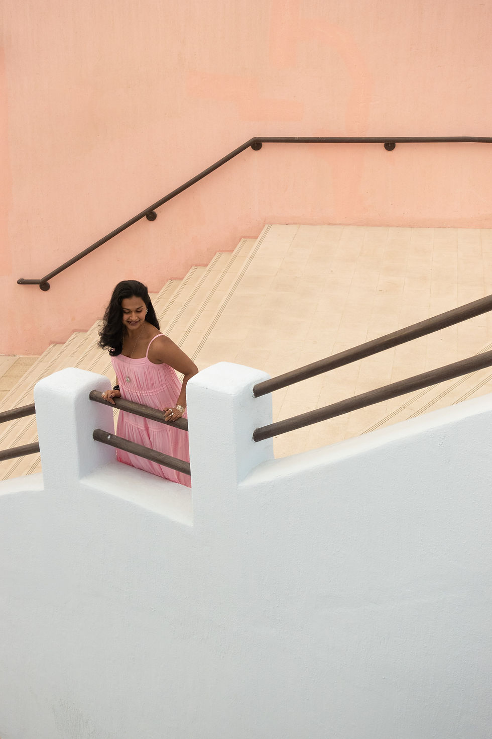 Woman in a pink dress smiles while descending pastel steps with dark railings against a peach wall, creating a serene mood.