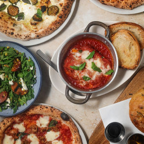 Overhead view of a rustic Italian spread featuring wood-fired pizza, fresh pasta, antipasti, and glasses of red wine on a worn wooden table, styled with olive branches, parmesan shavings, and linen napkins for a warm, inviting feel.