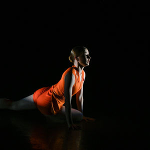 Robyn Pilecki is in an orange leotard and shorts. Her hair is in a low bun. She is posed on the ground, right leg bent and arms on either side. She is looking at the downstage left corner of the stage. She is the only thing lit in the photograph with a black background, creating a striking contrast.