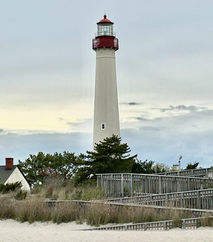 Tall white Cape May lighthouse with red top, sandy beach, and wooden boardwalk, a tourist attraction nearby.