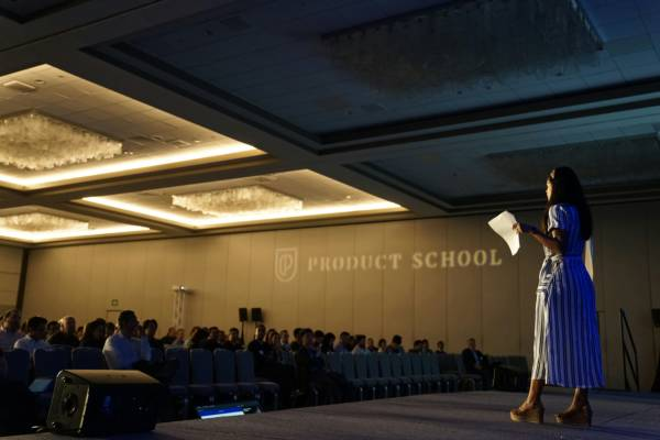 Woman speaks on a stage at a Product School event, holding papers before an audience in a dim conference hall.