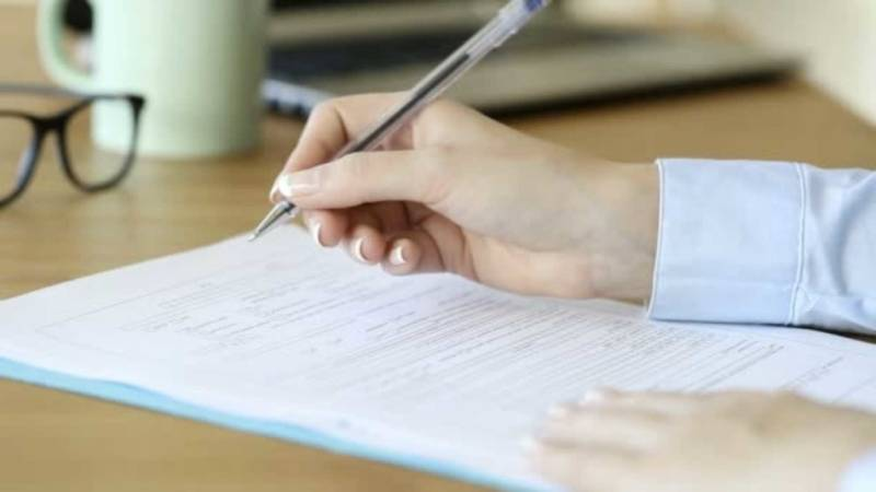 Hand filling out a form with a pen at a desk, with a mug and glasses blurred in the background
