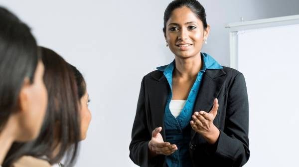 Smiling woman in black blazer presents to a small group in a bright office, gesturing as she speaks.