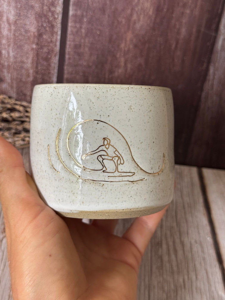 White speckled clay mug with gold linedrawn image of man surfing a wave