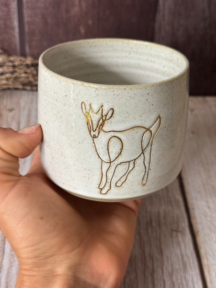 Hand holds a white speckled clay hug mug stamped with a gold-lined goat design