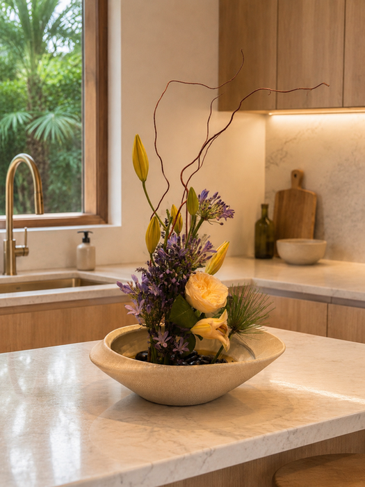Modern floral arrangement with white roses, purple blooms, sculptural branches, and tropical greenery styled in a ceramic bowl on a contemporary luxury kitchen island.