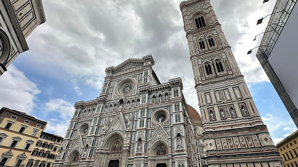 Ornate cathedral facade and bell tower with patterned white, green, and pink marble under a cloudy sky. Urban setting with nearby buildings.