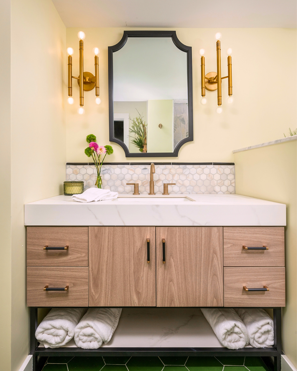 Designer bathroom by Prefixe Design showing a wood floating vanity with a centered mirror and brass sconces at eye level, illustrating correct vanity, mirror, and lighting heights