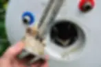 Hand holding corroded heating element removed from a water heater with blue and red valves; signs of mineral buildup. Background is blurred.