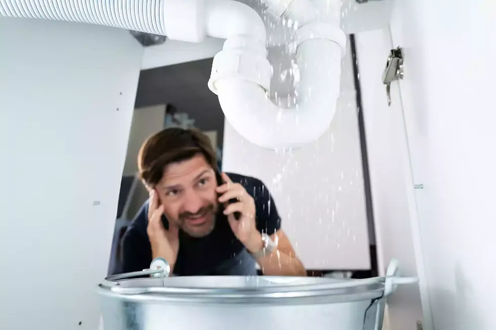 Man on phone under a leaking sink pipe, looking concerned. Water is splashing into a metal bucket below. Indoor setting, white background.