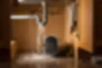 Leaking kitchen pipe under sink with water pouring into a tilted bucket, soaking the floor. Wooden cabinet background, wet towel hanging.