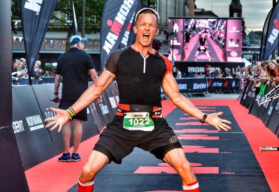 Runner celebrating at Ironman finish line, arms raised, wearing black and red attire with bib 1022. Crowd and banners in background. Excited mood.