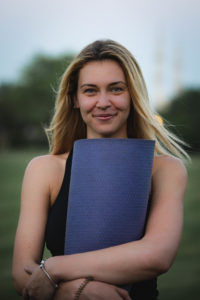 Woman smiling, holding a blue yoga mat in a grassy park. She's wearing a black top, with trees blurred in the background, conveying a serene mood.