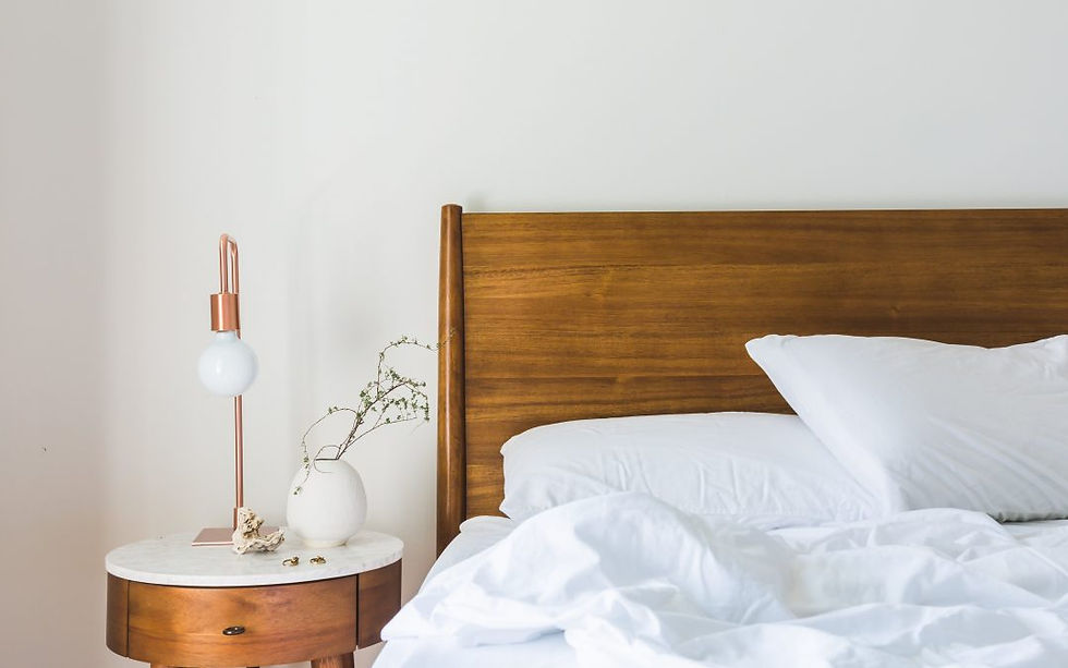 Cozy bedroom with wooden headboard, white pillows, and sheets. A round table hosts a lamp, vase with branches, and a calm mood.