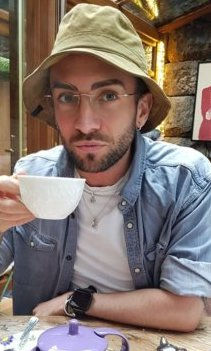A person wearing a hat and glasses sips from a white cup in a cozy cafe. Stone wall and wooden accents in the background. Relaxed mood.