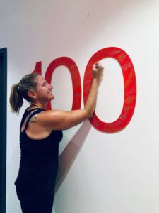 A woman in a black tank top happily writes on a large red "100" on a white wall, inside a room with a blue door frame.