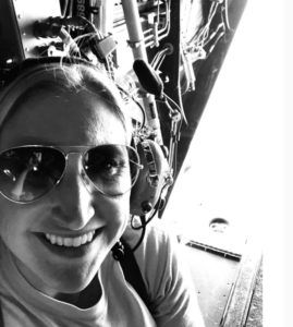 Person wearing aviator sunglasses and headphones smiles inside an aircraft cockpit. Cables and controls are visible; appears bright and cheerful.