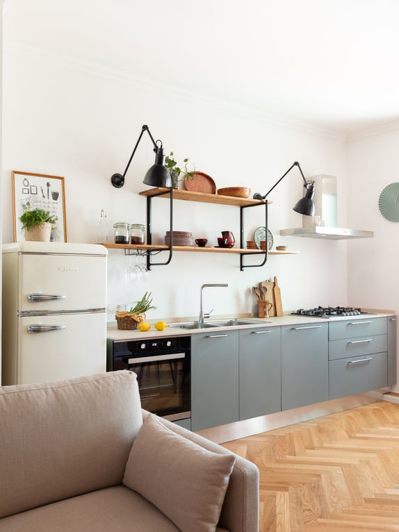 foto con vista sulla cucina in linea color verde salvia con frigo a libera installazione e a terra listoni in rovere posati a spina italiana. si intravede il divano beige.
