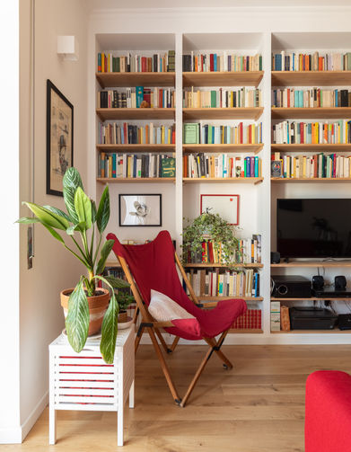 Vista stretta sul living. In primo piano la poltrona rossa, un comodino con una pianta e dietro la libreria in muratura con le mensole in legno, piena di libri. A terra il colore caldo del parquet a listoni. Pareti verniciate di bianco.