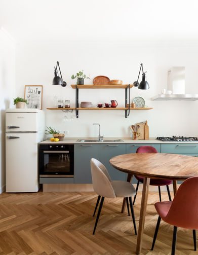 foto con vista sulla cucina in linea color verde salvia, il frigo è a libera installazione bianco. sopra la cucina ci sono due mensole con due appliques a braccio nere. tavolo in legno rotondo con sedie imbottite colorate. parquet posato a terra in rovere a spina italiana.