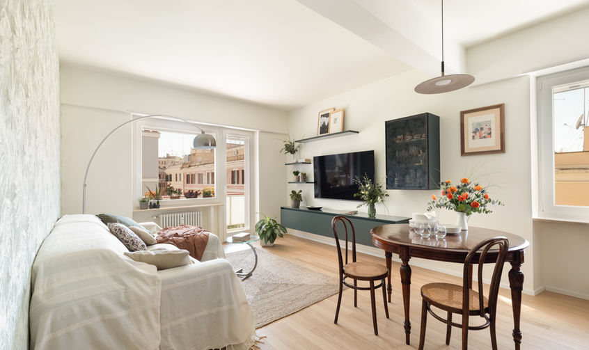 foto con vista su living dell'appartamento. divano a sinistra con rivestimento in tessuto bianco. in primo piano tavolo con sedie in legno e paglia di vienna. finestra che da su balcone sul fondo.