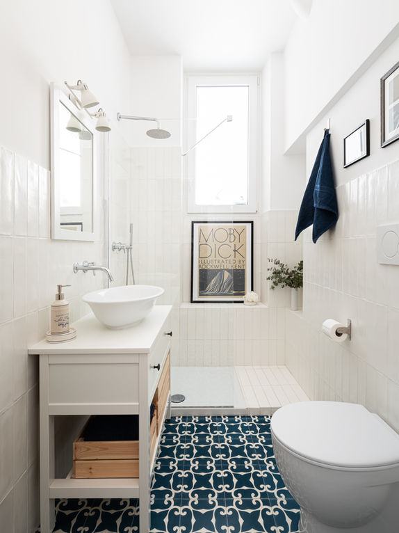 Foto del bagno. Le mattonelle bianche della parete sono state posate per il verso della lunghezza per slanciare l'ambiente, a terra cementine decorate sui toni blu e bianchi. Mobile lavabo in legno verniciato bianco con lavabo da appoggio in ceramica. Sanitari bianchi e rubinetteria cromata. Doccia con vetro walk-in e finestra alta.
