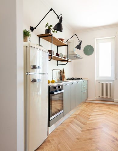 foto con vista sulla cucina che si trova a vista sul living. il frigo è a libera installazione e la cucina è sormontata da due mensole con due applique nere a braccio che illuminano il top. il pavimento è in listelli di rovere posati a spina italiana.