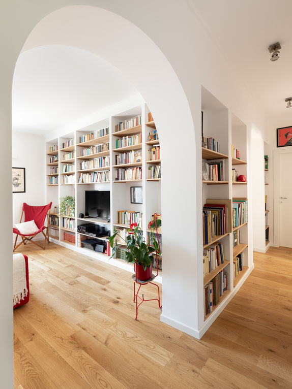 Vista dell'appartamento dall'ingresso. Si vede il corridoio sulla destra nel quale prosegue il sistema di libreria in muratura con le mensole in legno presente anche nel living. Listoni in parquet a terra. Toni caldi e morbidi.