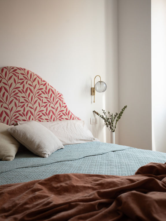 particolare con vista su letot matrimoniale composto da testiera letto realizzata su disegno con rivestimento in stoffa inglese con motivo vegetale color cremisi su fondo beige. applique con paralume in vetro a forma di globo e braccio color ottone. 