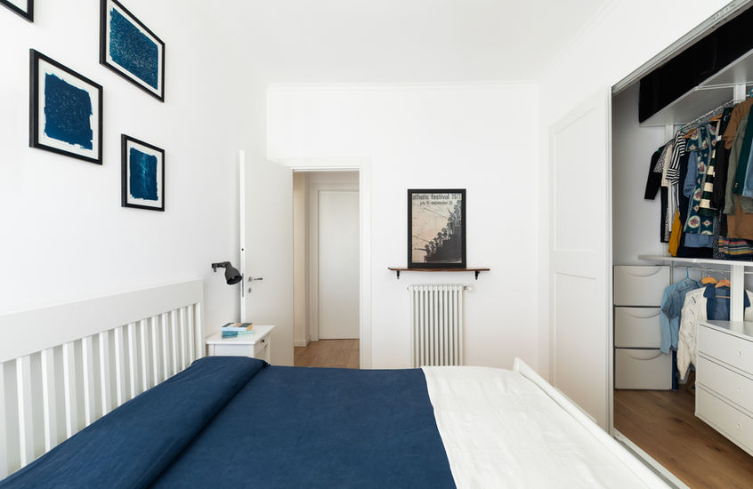 Vista della camera da letto. A destra cabina armadio aperta con vestiti appesi. Copriletto blu e pareti bianche