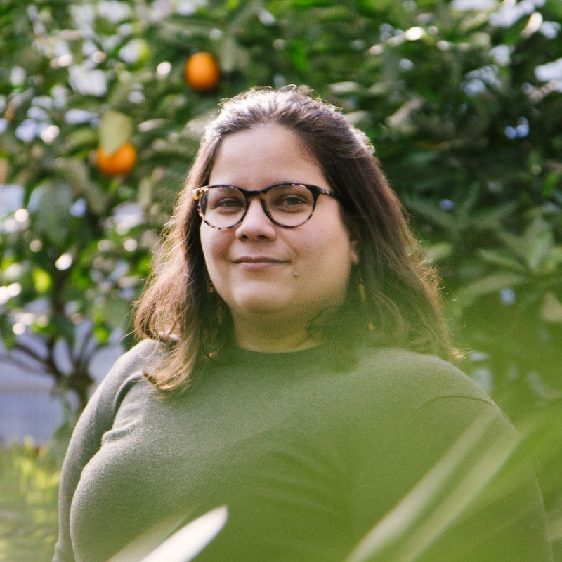 Gabriela Pereyra wearing glasses smiling in front of orange trees and leaves.