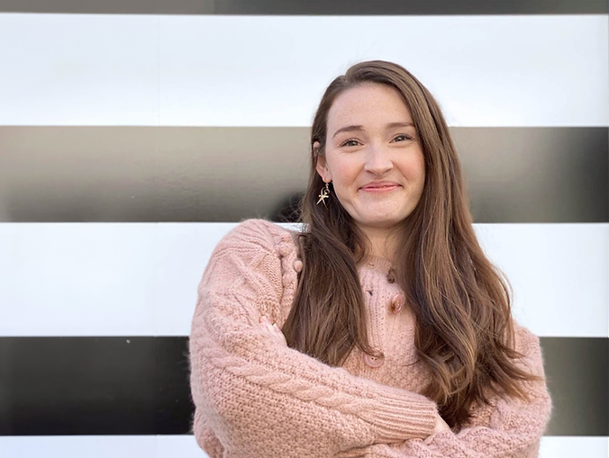 A Headshot of Alexandra Smith. She is wearing a light pink sweater and stands smiling with arms crossed in front of a black and white background.