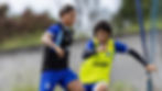 Dois jogadores do CSA disputam a bola durante treinamento no CT Gustavo Paiva. O atleta à esquerda conduz a bola enquanto é pressionado pelo companheiro que veste colete amarelo. A atividade acontece em campo de grama natural, com foco em trabalho de intensidade e marcação, tendo a trave e a vegetação ao fundo.