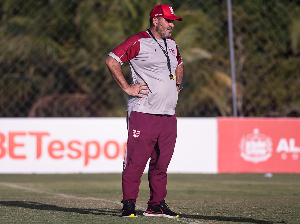 Treinador do CRB observa o treino à beira do gramado, uniforme vermelho e cinza, com fundo de campo e escudo do clube ao fundo.