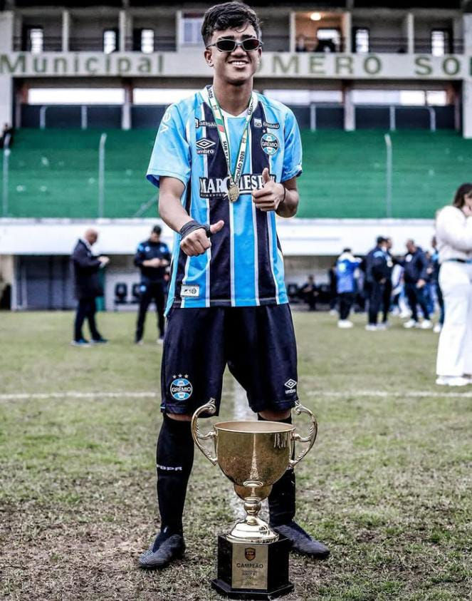 Benjamin Santos posa sorridente com óculos escuros e medalha no pescoço, vestindo o uniforme do Grêmio, ao lado de um grande troféu de campeão em campo gramado; ao fundo, o Estádio Municipal Homero Soldatelli e pessoas comemorando o título.