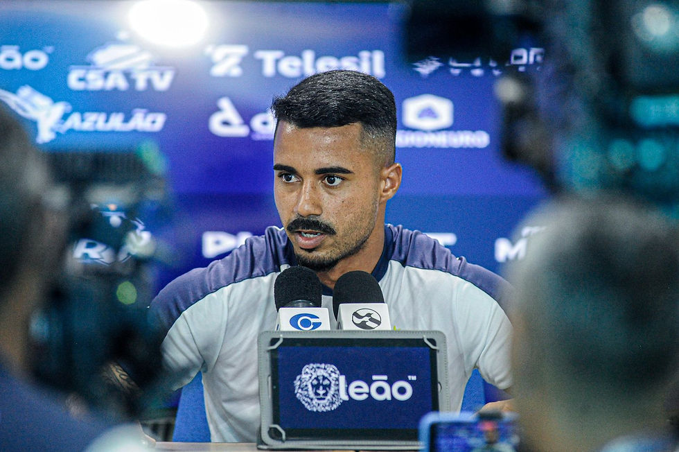 Fotografia em close do volante Fabrício Bigode falando ao microfone durante coletiva de imprensa no Centro de Treinamento do CSA. Ele veste uniforme branco e azul, e ao fundo aparecem logos de marcas como Telesil e CSA TV.