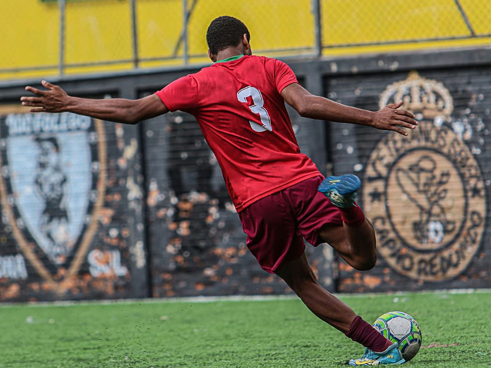 Adryan Gabriel, jovem zagueiro e volante de 15 anos, veste uniforme vermelho com o número 3 nas costas enquanto faz um passe com a perna direita em campo sintético. A imagem capta o momento exato da batida na bola, com os braços abertos para equilíbrio, mostrando técnica e concentração.