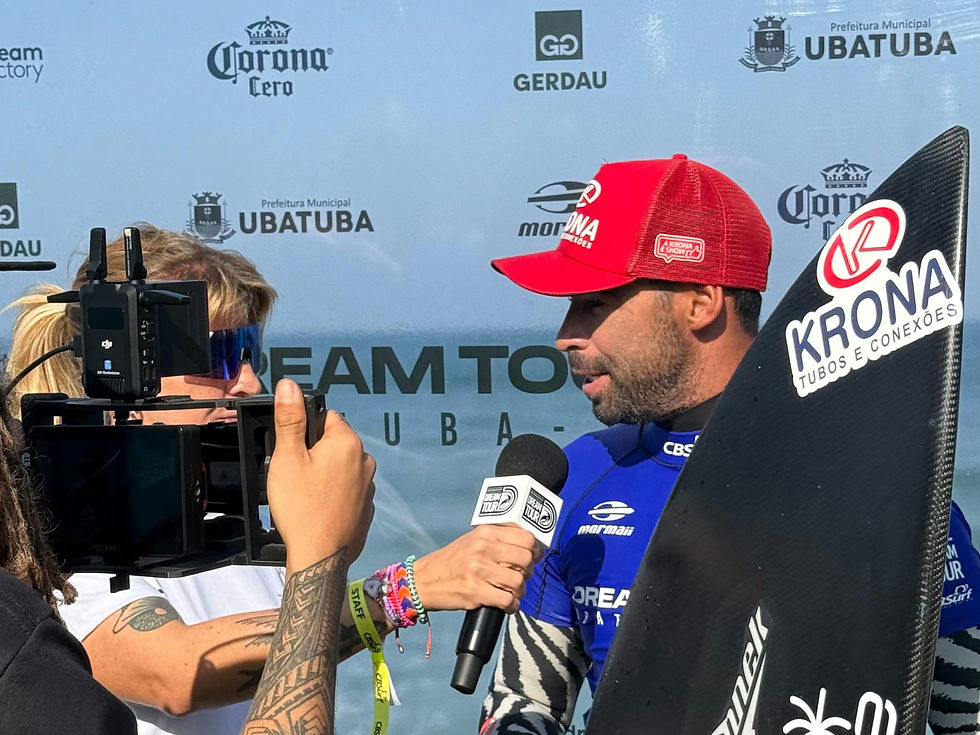 Surfista Amando Tenório concede entrevista segurando sua prancha após vencer bateria no Dream Tour em Ubatuba. Ele veste camisa azul do campeonato, boné vermelho da Krona e aparece diante do backdrop oficial do evento, com logos de patrocinadores e da Prefeitura de Ubatuba ao fundo.